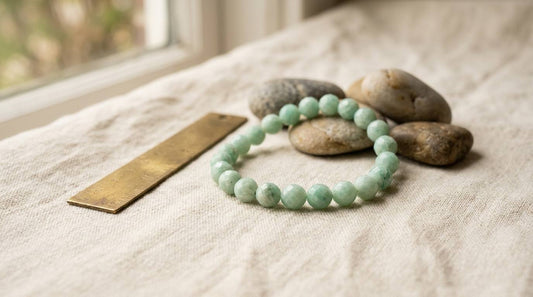 Pale green gemstone bracelet beside a brass ruler and river stones on cream linen